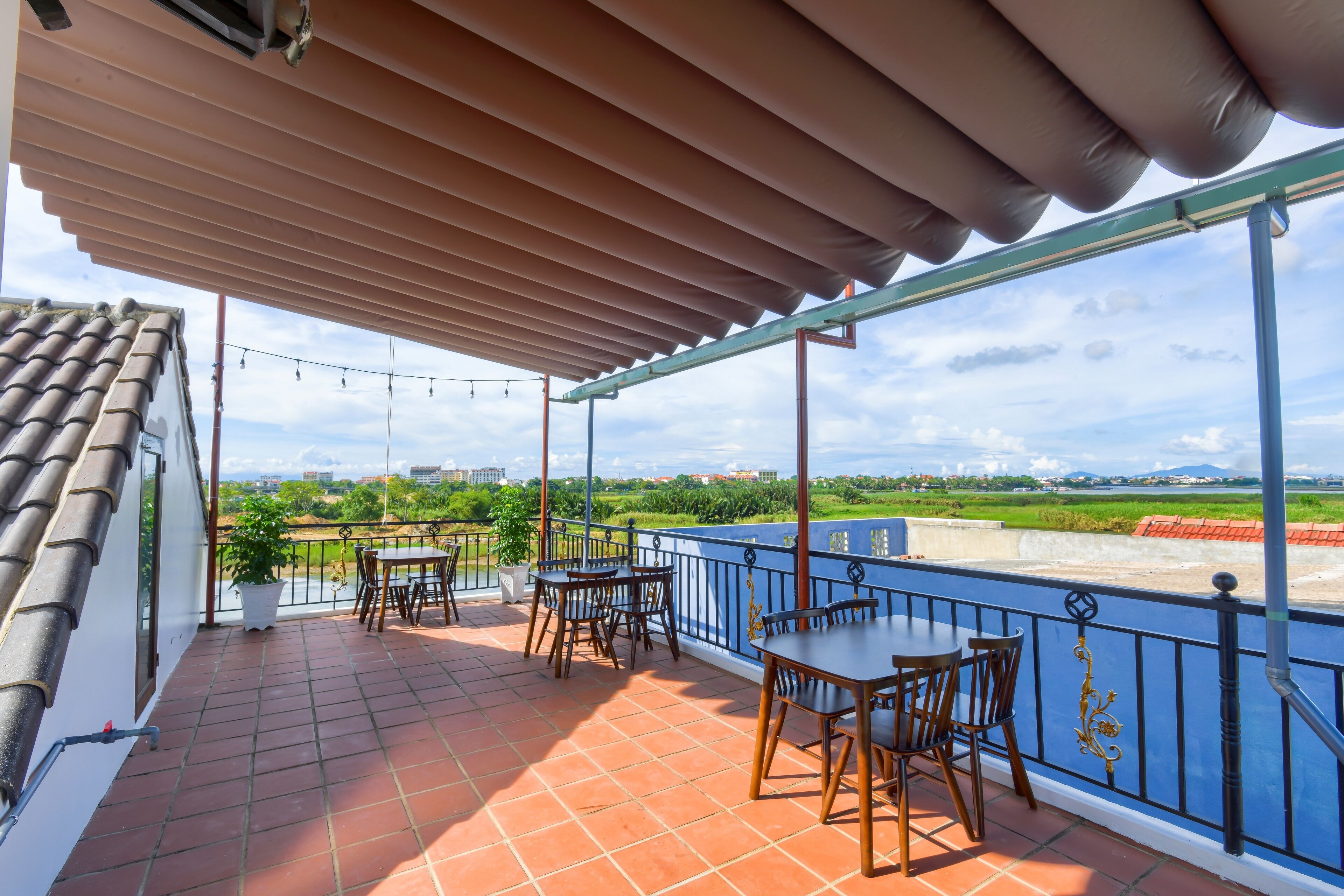 A terrace with tiled flooring, wooden tables and chairs, a blue railing, and a view of a green landscape and buildings in the distance.