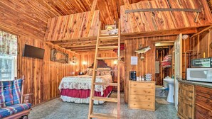 Interior de una cabaña de madera con cama en altillo, escalera, televisor, baño y comedor.