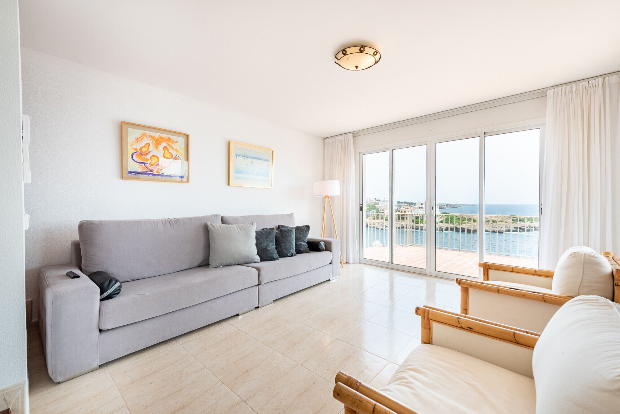 Een moderne woonkamer met een grijze bank, een vloerlamp en uitzicht op zee door grote ramen.