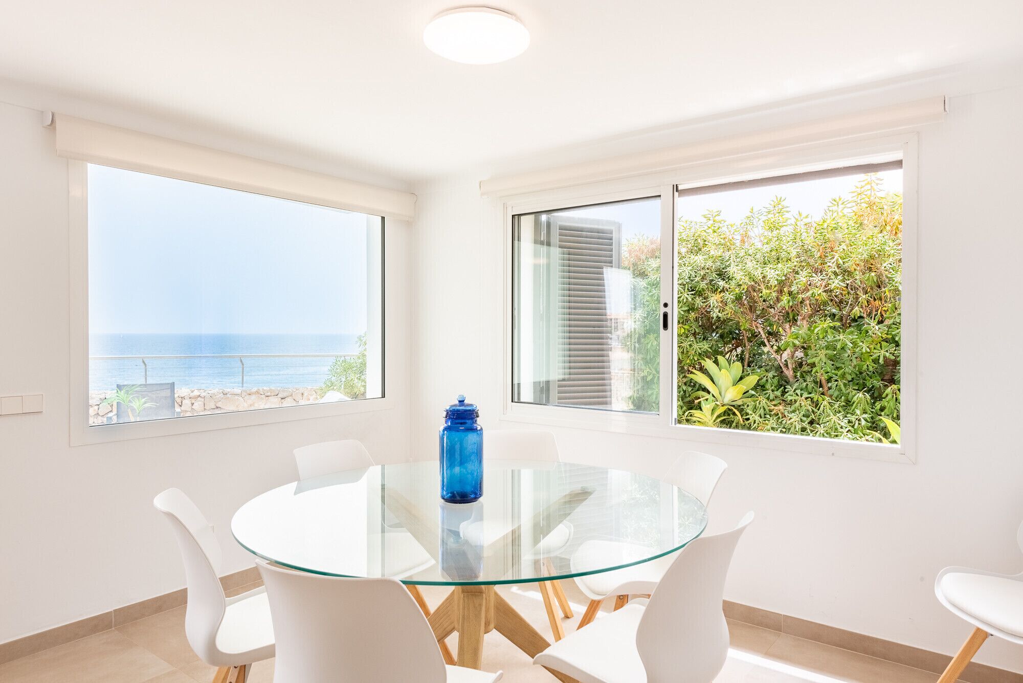 Een kamer met een glazen tafel, witte stoelen, een blauwe waterfles en grote ramen met uitzicht op zee en begroeiing.