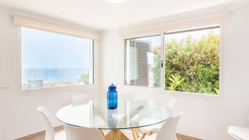 Een kamer met een glazen tafel, witte stoelen, een blauwe waterfles en grote ramen met uitzicht op zee en begroeiing.