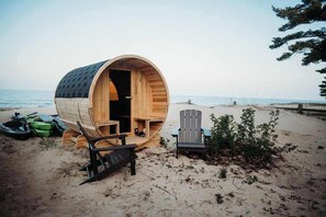 Une structure en forme de tonneau, faite de bois, se trouve sur une plage et contient une chaise et un banc à l’intérieur.