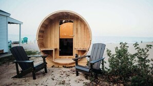 A wooden barrel structure with a door, two Adirondack chairs, and a small porch area on a beach.