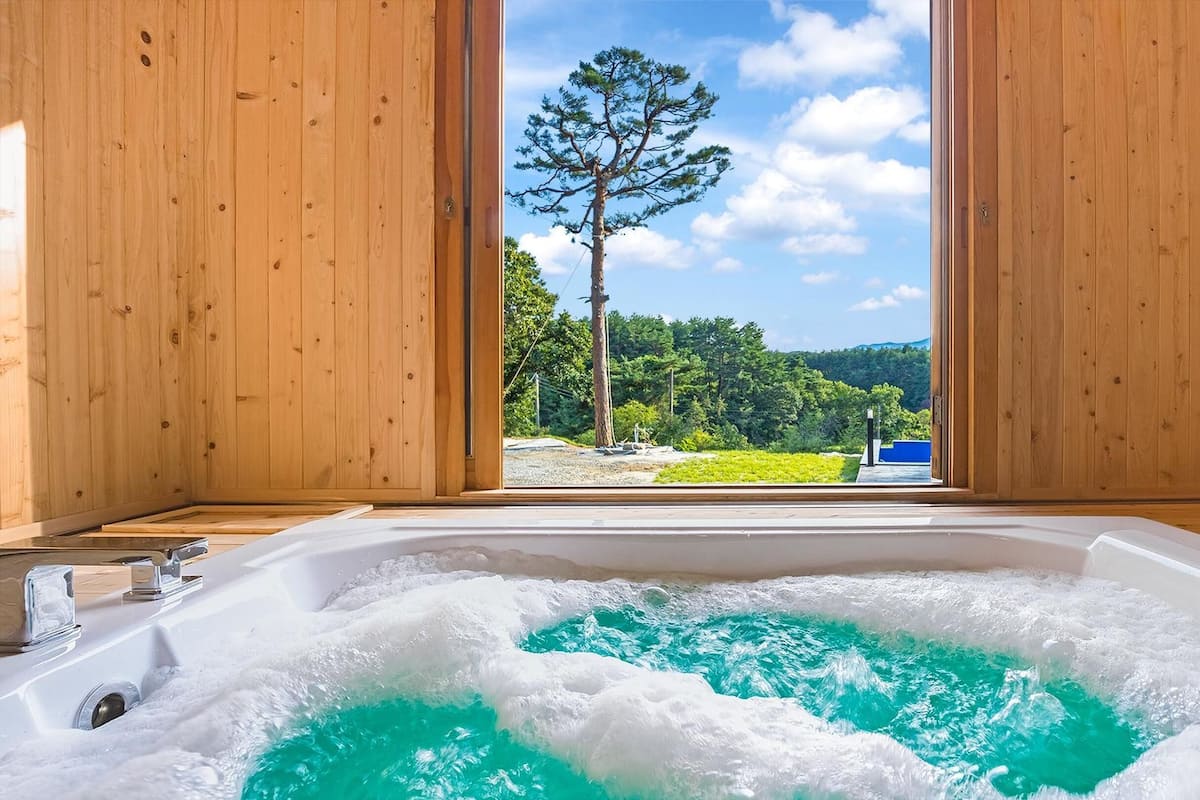 Una habitación de madera con un ventanal grande, una bañera llena de agua y vistas a un árbol y al paisaje.