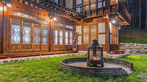 A traditional wooden house with large windows and a stone pathway leading to a circular fire pit area.
