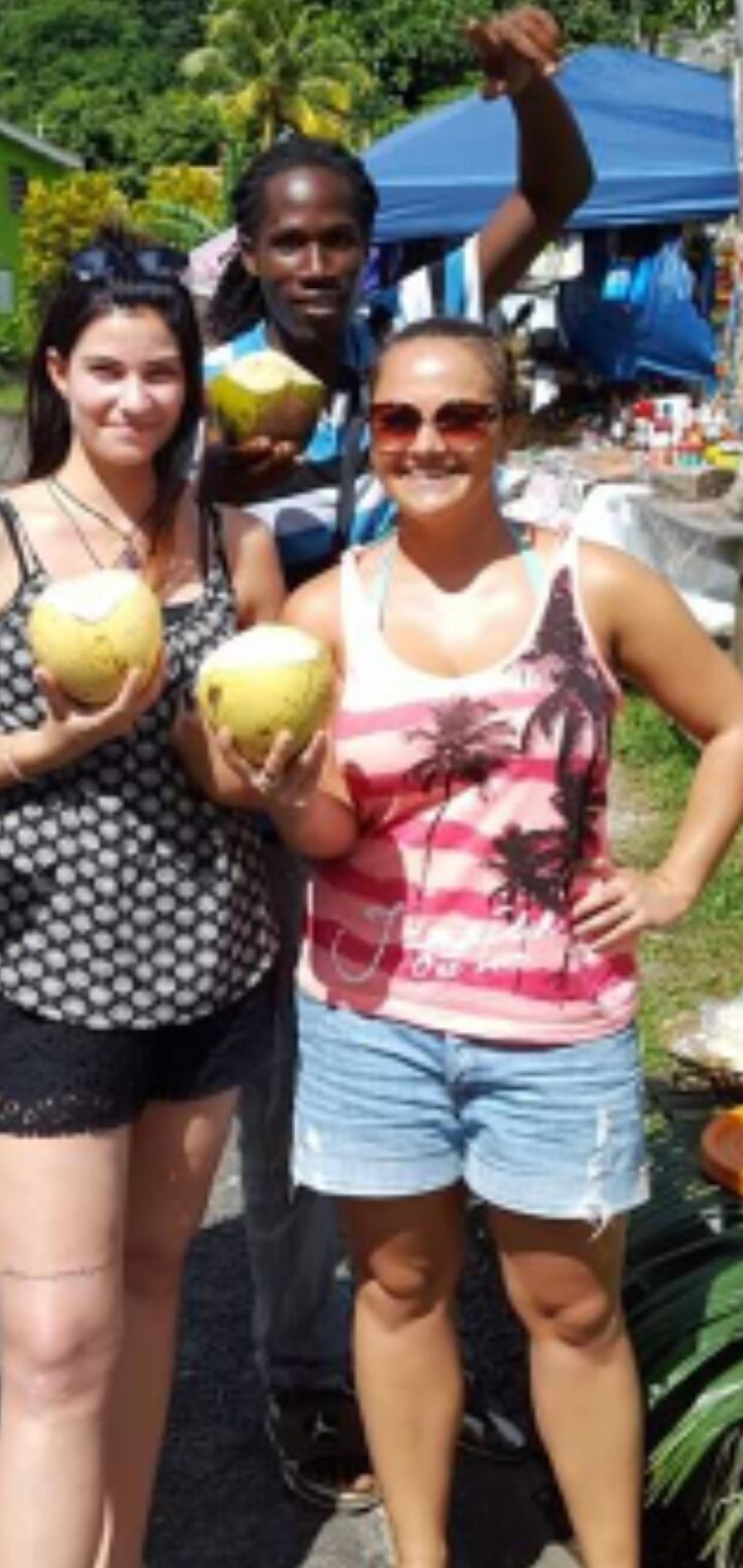 A group of people outdoors, some holding coconuts, with a blue tent and greenery in the background.
