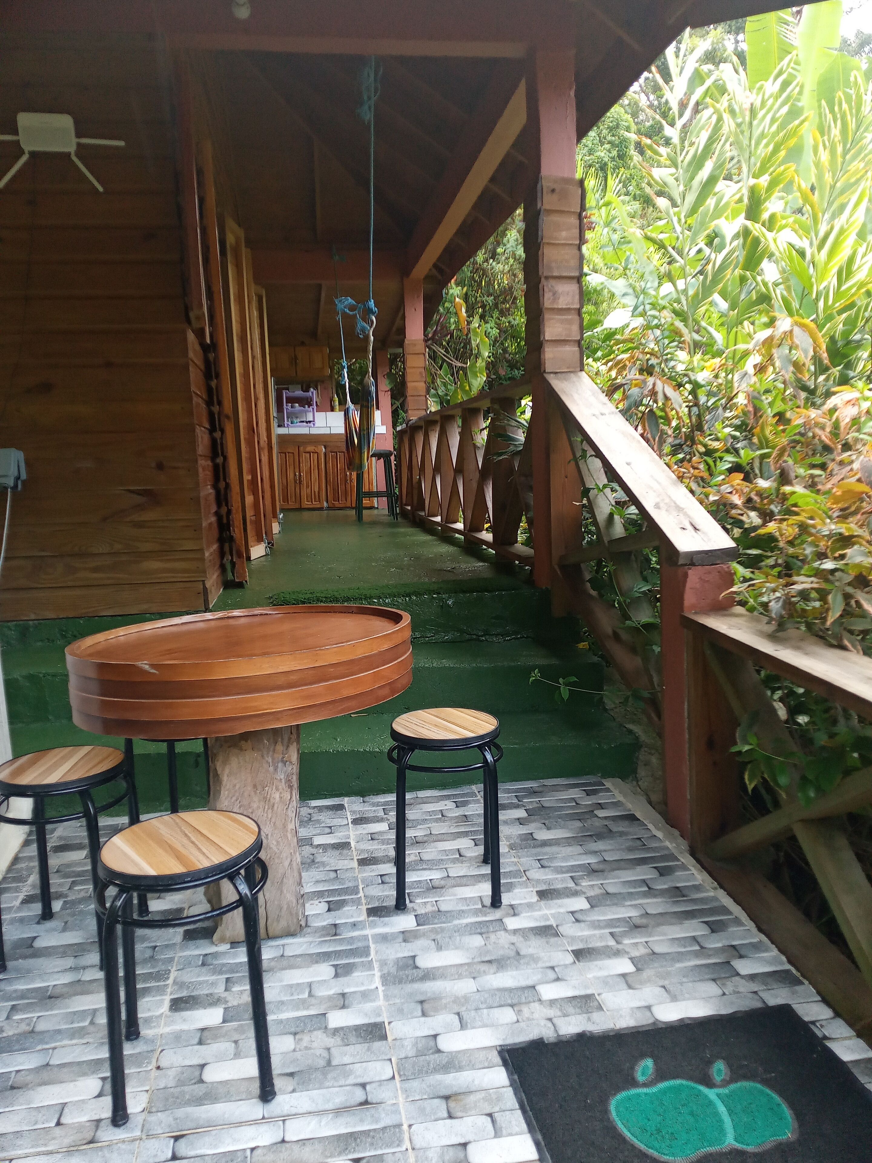 A covered outdoor seating area with a wooden table and stools, a brick floor, and a green mat with a fruit design.
