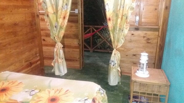 A wooden room with a bed, curtains, a small table with a lantern, and a door leading to an outdoor area. - Berkey's Jungle Paradise (Roseau)