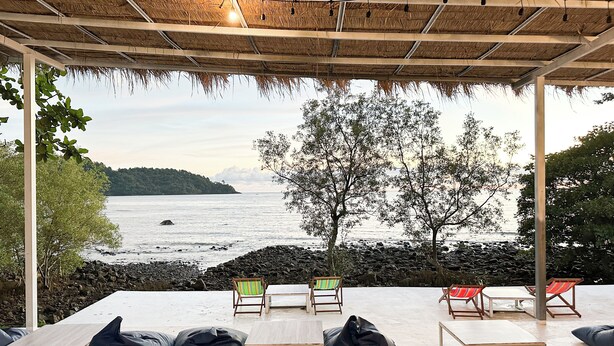Un área de descanso frente a la playa, con vista al mar, techo de paja y un árbol al fondo.