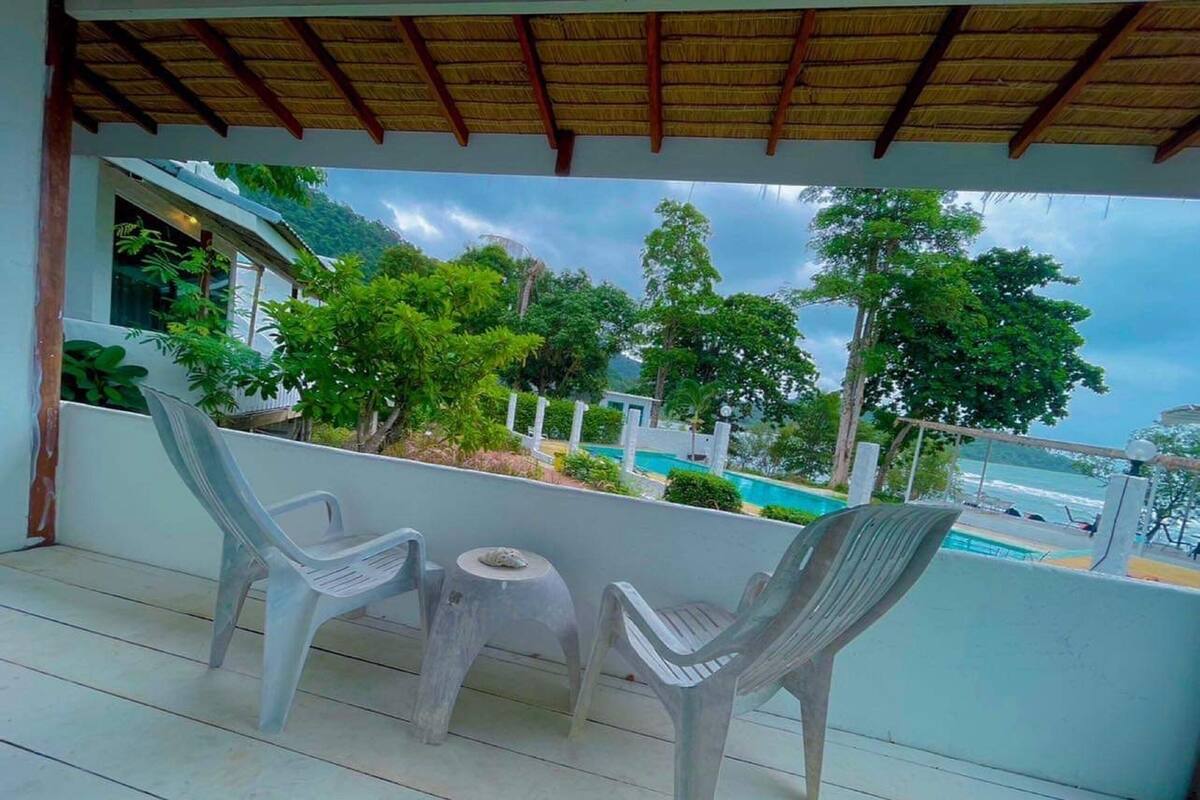 A covered outdoor seating area with two plastic chairs and a small round table, overlooking a pool and lush greenery.