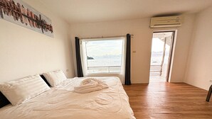 Una cama bien hecha con una toalla, un cuadro de un paisaje urbano en la pared y vista al mar por la ventana.