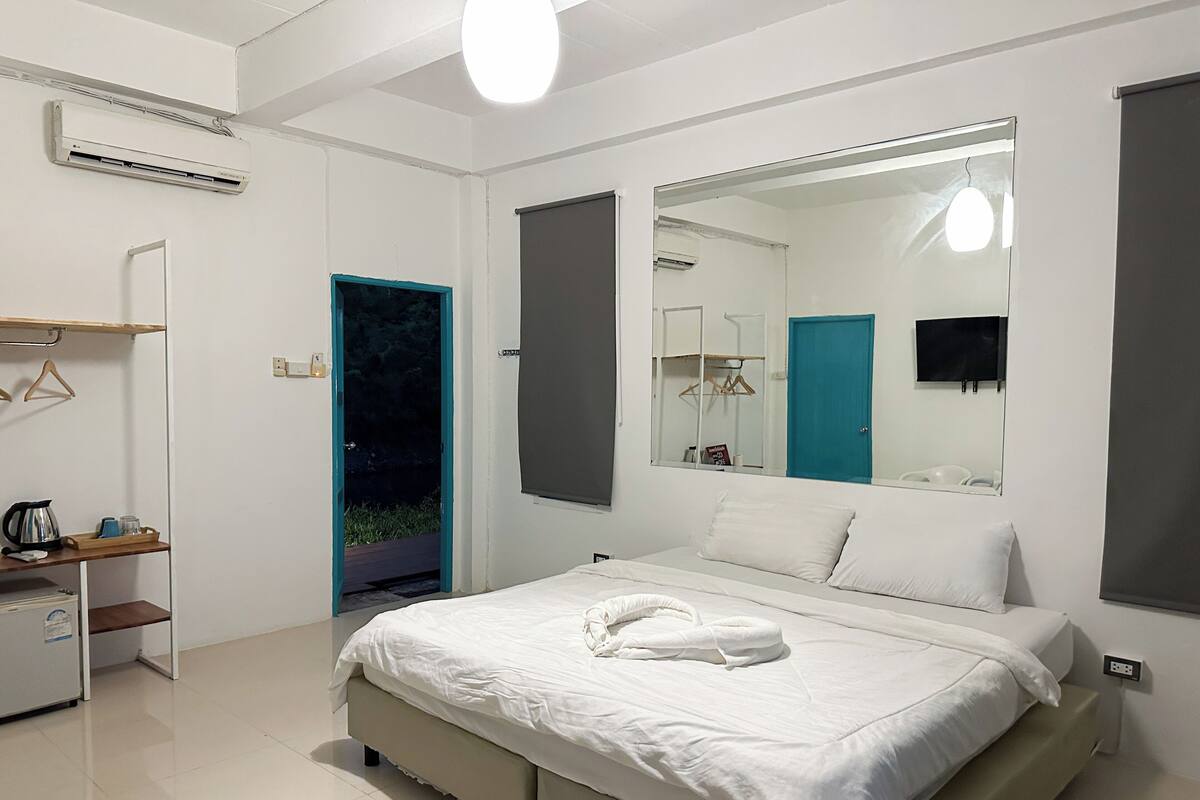 A modern bedroom with a large bed, a bedside table with a kettle, a mirror, and a view of a blue door.
