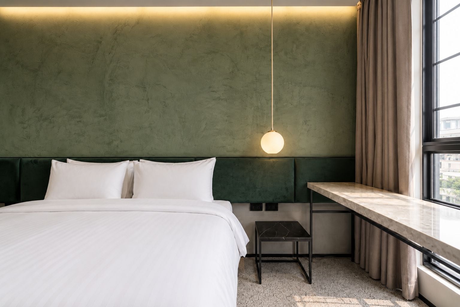 Une chambre d’hôtel avec un lit, un bureau, une chaise, un luminaire suspendu et une fenêtre avec des rideaux.