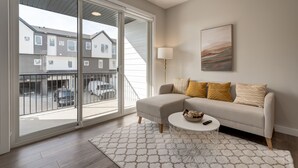 A modern living room with a large sliding glass door leading to a balcony, a beige sofa with patterned cushions, a round coffee table, and a floor lamp. - *MODERN* 2BDRM Close to Airport & Major Highway (Calgary)