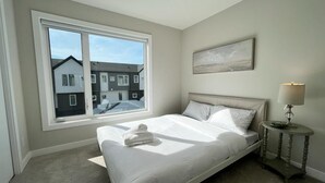 A bedroom with a large window, a bed, a bedside table with a lamp, and a painting on the wall. - *MODERN* 2BDRM Close to Airport & Major Highway (Calgary)