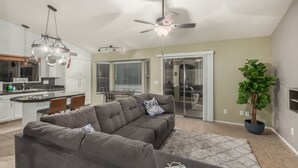 Una sala moderna con un sofá modular gris, un ventilador de techo y una cocina al fondo.