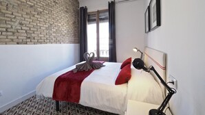 Ein Hotelzimmer mit Ziegelwand, einem Bett mit weißer Bettwäsche und roten Kissen, einer roten Tagesdecke, einer Schreibtischlampe und einem Fenster mit Vorhängen.