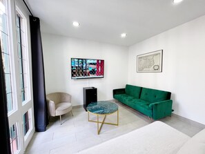 Un salon moderne avec un canapé vert, une table basse ronde, un fauteuil beige, un téléviseur fixé au mur et une carte encadrée.