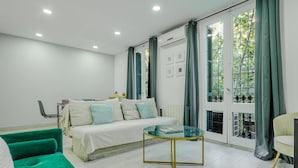 Una habitación moderna con una cama, un sofá verde, una mesa de centro de vidrio y ventanas amplias con cortinas.