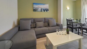 A modern living room with a grey sectional sofa, a glass coffee table with wine glasses and a bottle, and a painting of a harbor scene on the wall. - Sagrada Familia Penthouse (Barcelona)