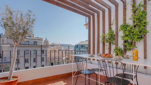 Een balkon met een plant in pot, een tafel met stoelen en een uitzicht op stadsgebouwen.