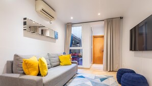 Ein modernes Wohnzimmer mit einem grauen Sofa, gelben Kissen, einem Teppich mit geometrischem Muster, einem blauen Sitzsack und einem an der Wand montierten Flachbildfernseher.