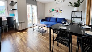 En moderne stue med en blå sofa, et spisebord dækket til to personer, og et køkkenområde med komfur og vindue med udsigt til en balkon.