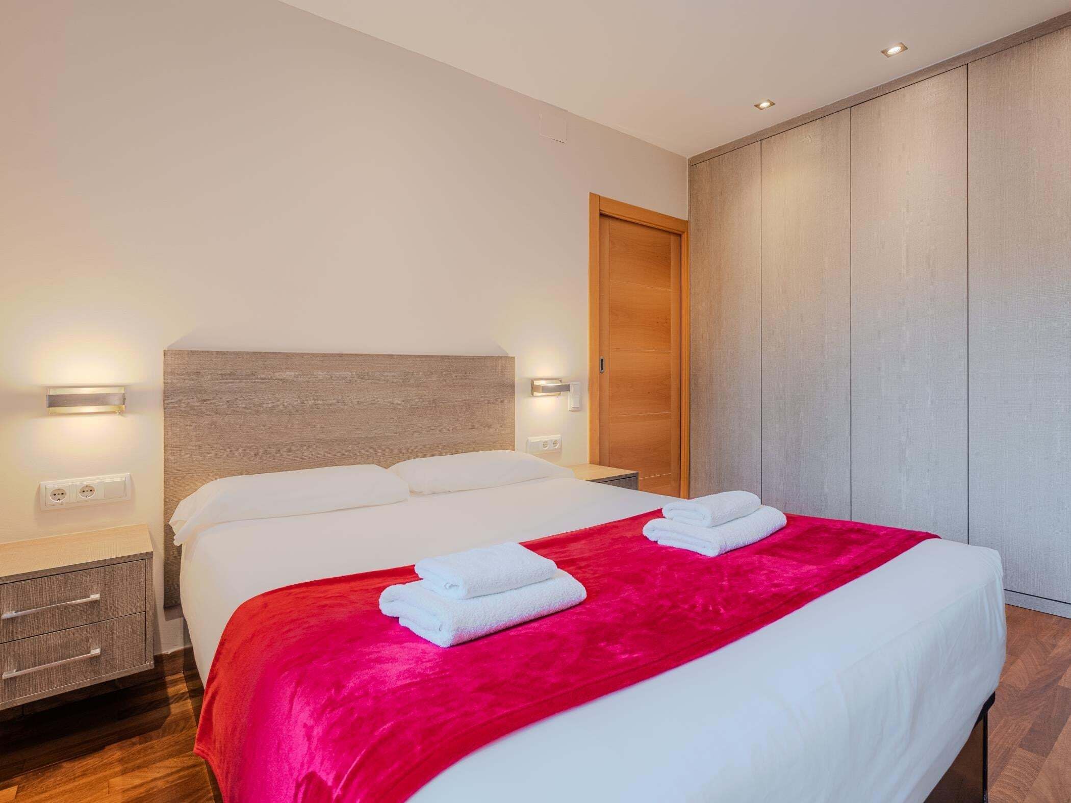 Une chambre d’hôtel moderne avec un grand lit, deux tables de chevet, une armoire en bois et un mur d’accent rouge.