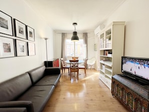 A living room with a black sofa, a dining table with chairs, a TV on a cabinet, and framed pictures on the wall.