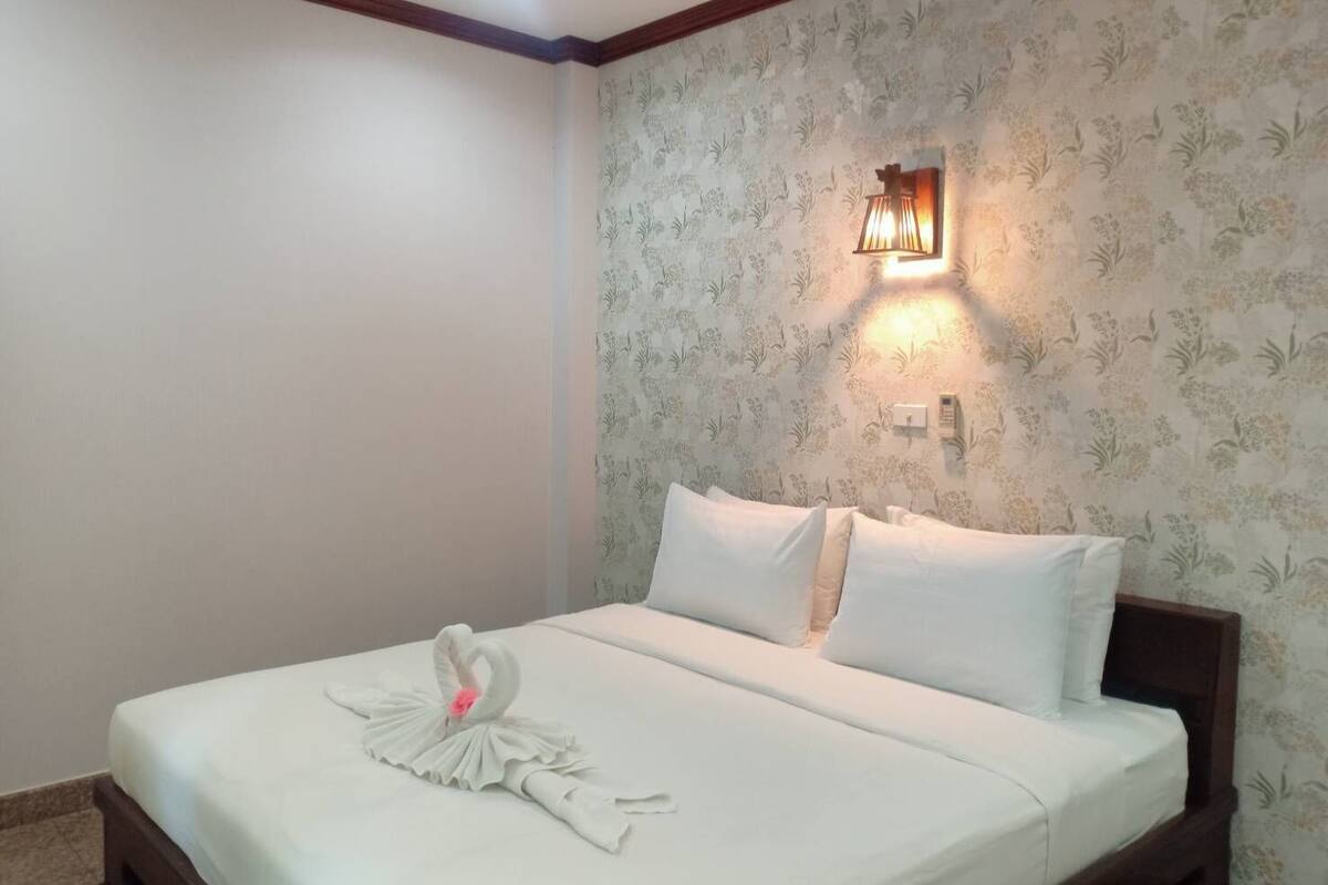 A neatly made bed with white linens and a swan-shaped towel decoration. A wall-mounted light fixture is visible on the wall.