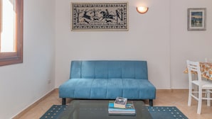 A living room with a blue sofa, a coffee table with books, a wall-mounted tapestry, and a dining area with a table and chairs. - La Dimora Di Montelittu With AC (Loiri Porto San Paolo)