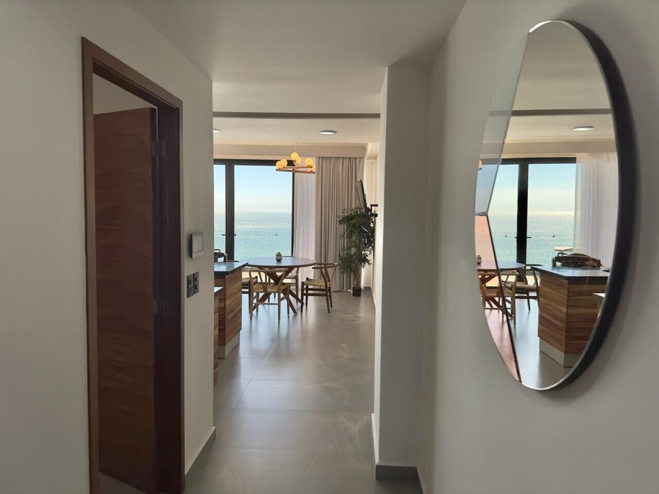 A modern hotel room with a large window, a dining area with a table and chairs, and a mirror on the wall.
