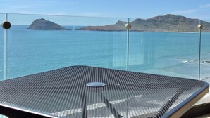 Een balkon met glazen hekwerk met uitzicht op een rustige zee en verre bergen.