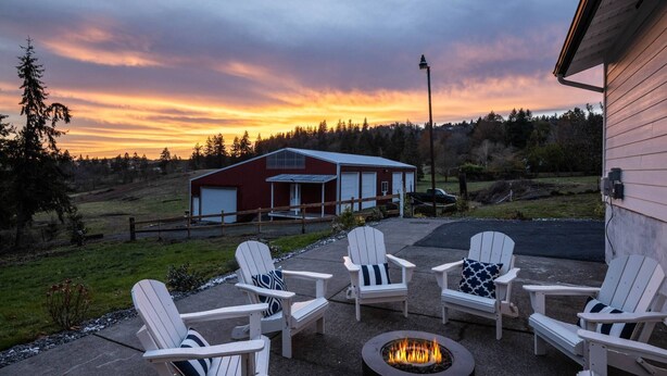 A patio with four Adirondack chairs and a fire pit, overlooking a red barn and a scenic sunset.