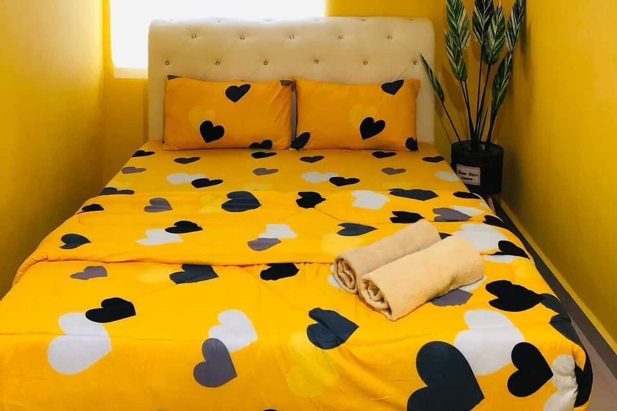 Uma cama com colcha amarela estampada com corações pretos e brancos, cabeceira branca e toalha enrolada sobre a cama.