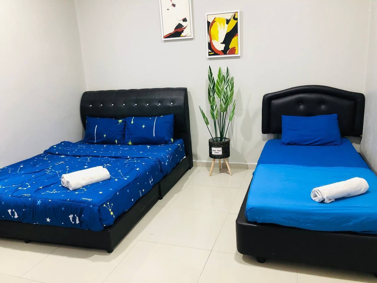 Two single beds with blue bedding and white towels, a potted plant, and two abstract paintings on the wall.
