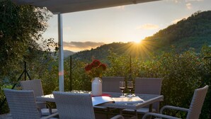 Un área de comedor al aire libre con sillas de mimbre y una mesa para dos, con un jarrón de flores rojas y una copa de vino.