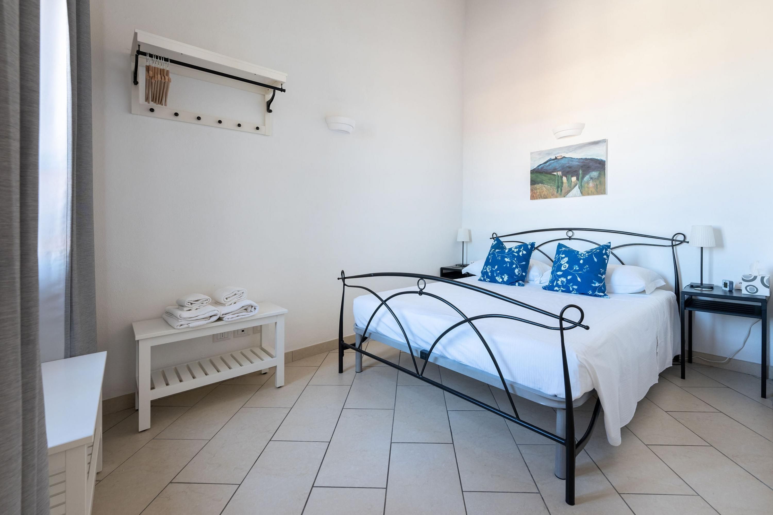 Een slaapkamer met een metalen bed, witte beddengoed, een nachtkastje met een lamp, een klein bankje en een wandplank met handdoeken.