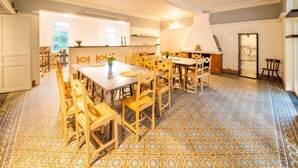 A spacious dining area with a large wooden table and chairs, a patterned floor, and a kitchen in the background.