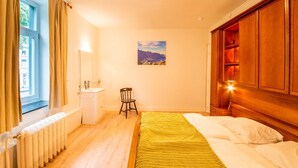 A bedroom with a wooden headboard, a bed with white linens, a radiator, a window with curtains, a small stool, a sink, and a painting on the wall.