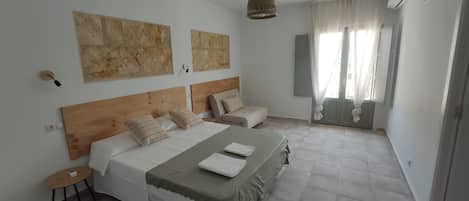 A bedroom with two beds, a wooden headboard, a bedside table, a hanging light fixture, and a view of the outdoors through a window with curtains.