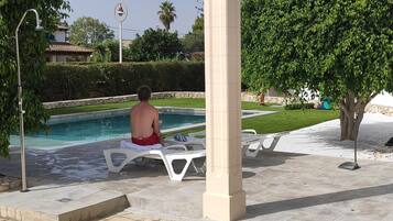 A pool area with a person sitting on a lounge chair, a large umbrella, and a stone pillar.