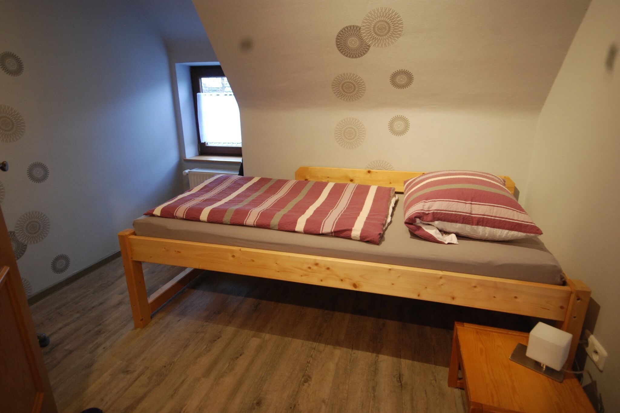 Una habitación pequeña con una cama individual de estructura de madera, una colcha a rayas y una almohada. Hay una mesita de noche de madera con una lámpara y un enchufe. La habitación tiene un techo inclinado con diseños decorativos en la pared y una ventana con persianas.