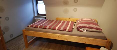 A small, single-bed room with a wooden frame, a striped bedspread, and a pillow. There is a small wooden nightstand with a lamp and a power outlet. The room has a sloped ceiling with decorative wall patterns and a window with blinds.