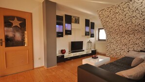 Ein modernes Wohnzimmer mit Holzboden, einem schwarzen Sofa, einem weißen Couchtisch, einem Fernseher auf einem schwarzen Schrank und einer Wand mit Muster.