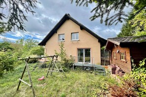 Une maison à deux étages avec un toit en pente, une terrasse en bois et un ensemble de jeux avec balançoire dans le jardin.