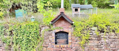 Un edificio de piedra rústico con una chimenea de ladrillo, rodeado de follaje y una pequeña construcción de madera al fondo.