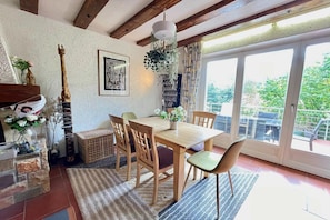 Un coin repas avec une table et des chaises en bois, une plante suspendue, une guitare et une fenêtre donnant sur un paysage verdoyant.