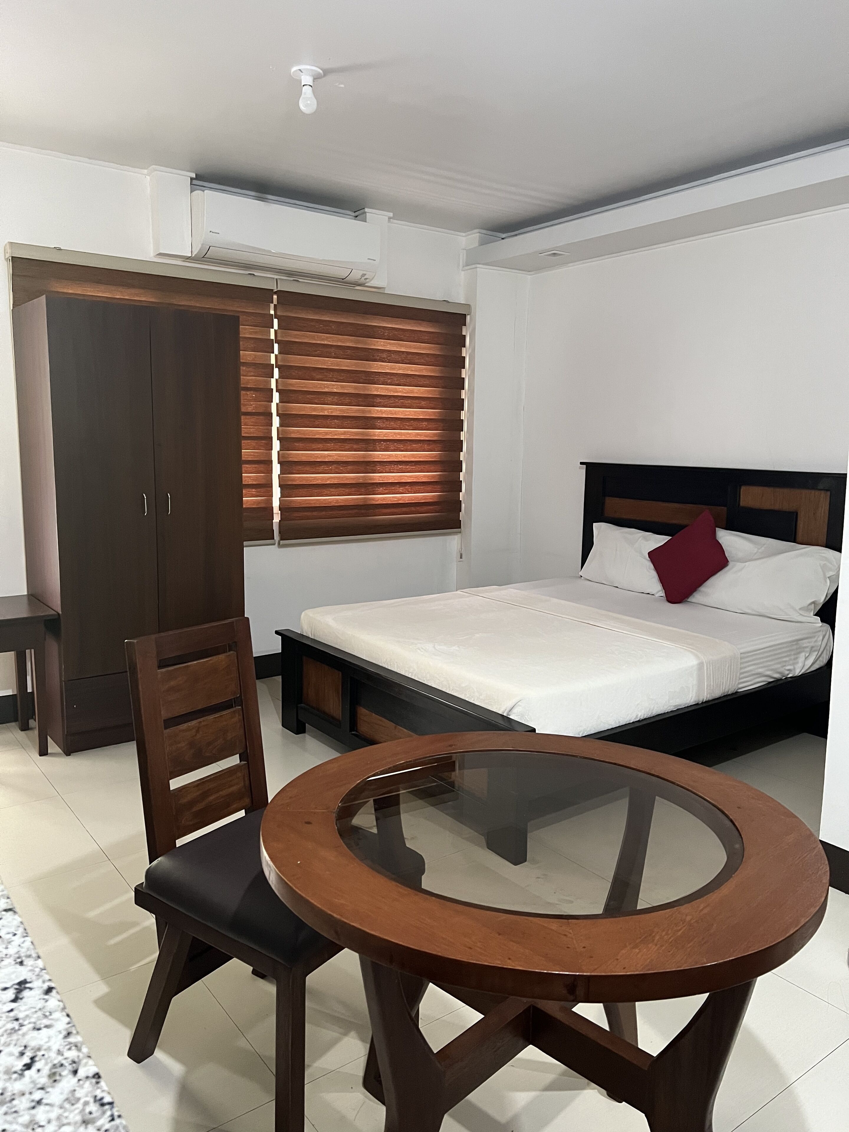 A bedroom with a bed, a wooden wardrobe, a round glass table, and a chair.