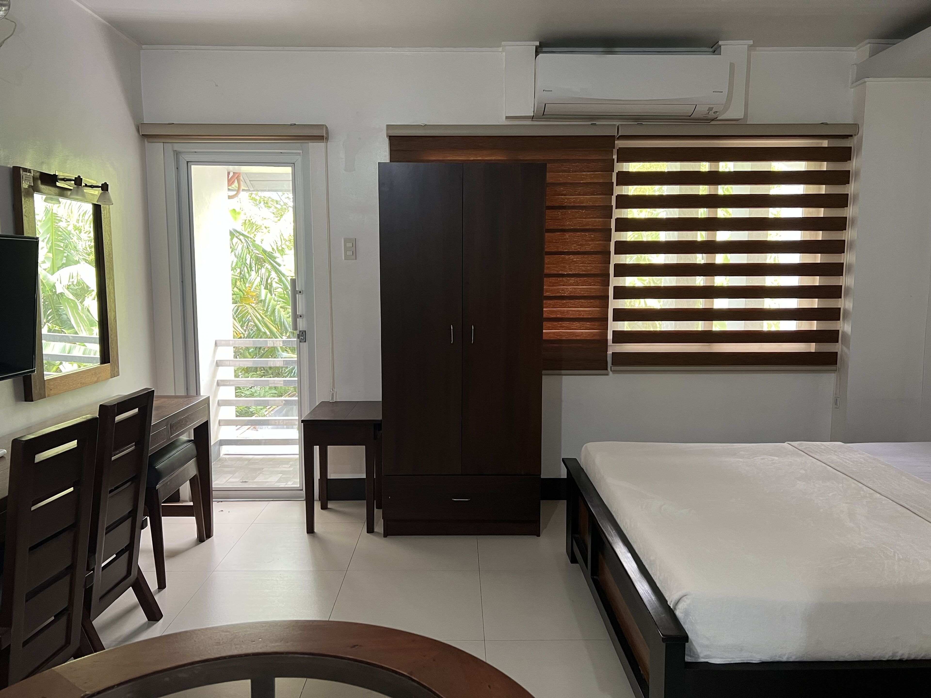 A bedroom with a bed, a desk with chairs, a wardrobe, and a window with blinds.